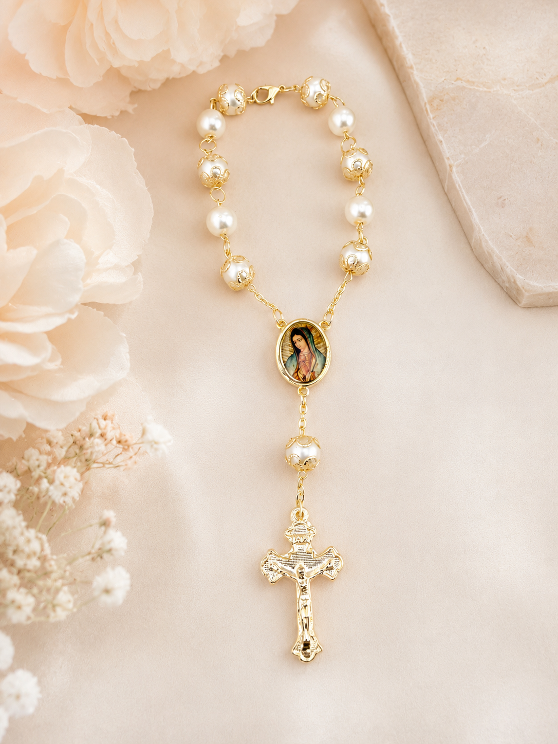 A gold and pearl rosary necklace featuring a central oval image of the Virgin Mary and a dangling crucifix, styled on a soft cream fabric with pale flowers.