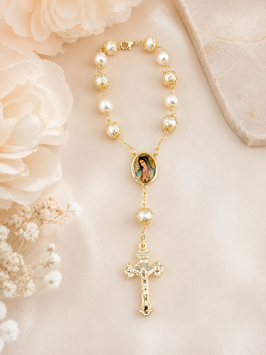 A gold and pearl rosary necklace featuring a central oval image of the Virgin Mary and a dangling crucifix, styled on a soft cream fabric with pale flowers.